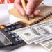 A woman's hand writing down the cash flow in a notebook with a pen, counting cash on a calculator